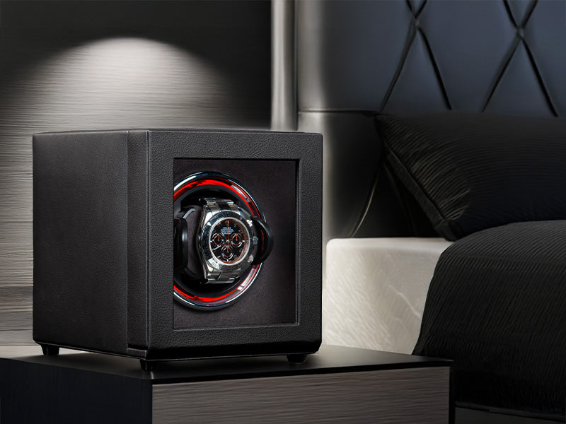 Black watch winder with a watch inside on a nightstand next to a bed.